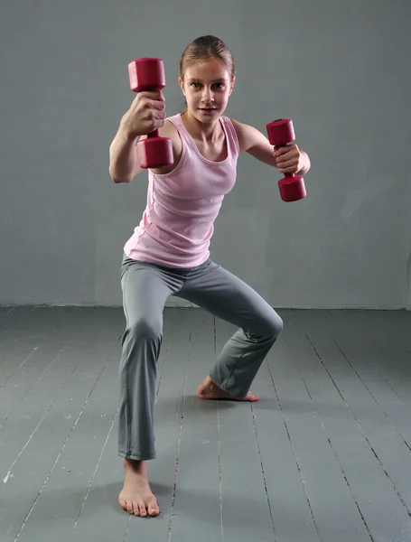 Teenage sportive girl is doing exercises with dumbbells to develop muscles on grey background. Sport healthy lifestyle concept. Sporty childhood. Teenager exercising with weights.