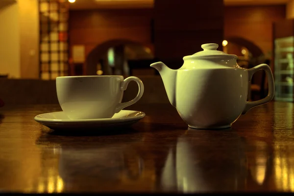 Tea time. White teacup and teapot on the wooden table in dark room, side view