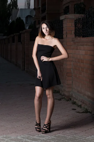 Cute brunette woman in short black dress is posing against red brick wall