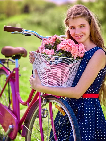 Girl wearing blue polka dots sundress rides bicycle with flowers basket.