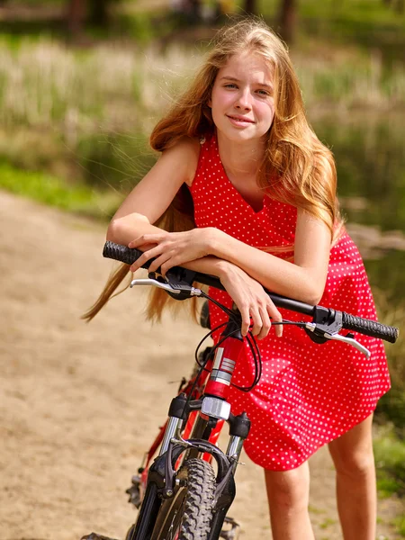 Girl wearing red polka dots dress rides bicycle into park.