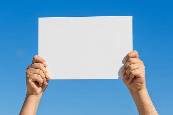 Blank white board in hands against blue sky background