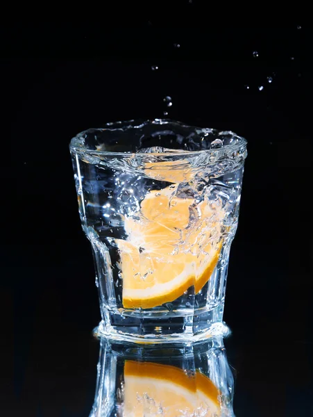 Slice of lemon splashing into a glass of water with a spray of water droplets in motion suspended in the air above the glass on a dark background.