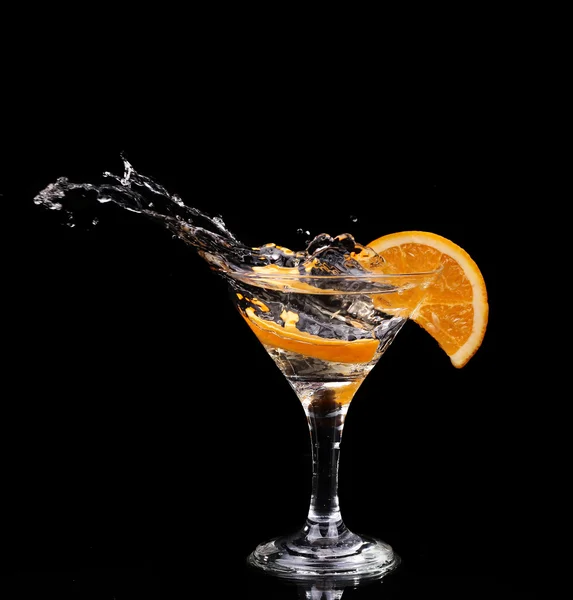 Slice of lemon splashing into a glass of water with a spray of water droplets in motion suspended in the air above the glass on a dark background.