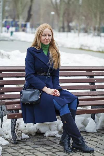 Young woman in a blue coat sitting on a bench in winter park
