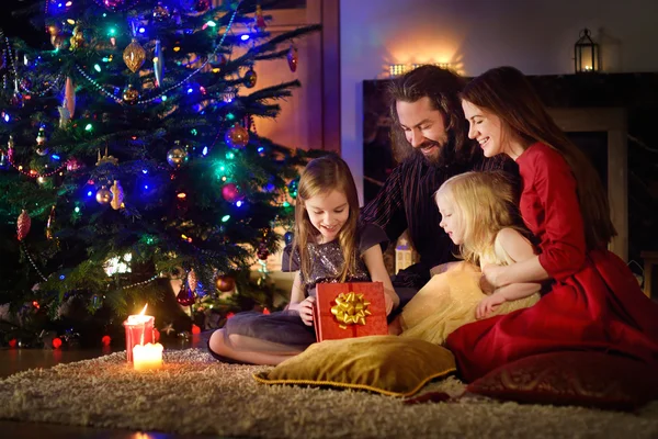 Young happy family of four unwrapping Christmas gifts by a fireplace