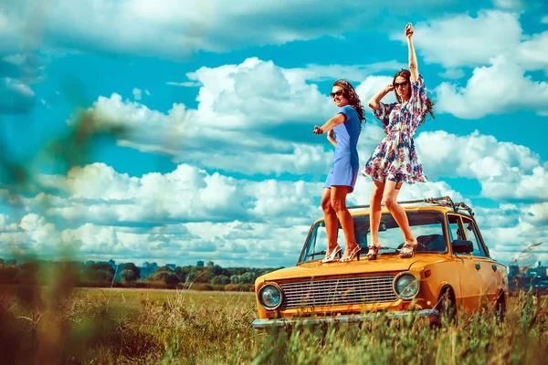 Two beautiful woman are dancing on the old car. Green fields on