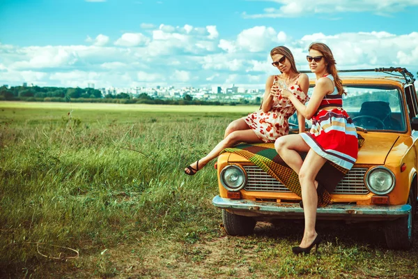 Two beautiful women on the picnic are drinking wine on the car.