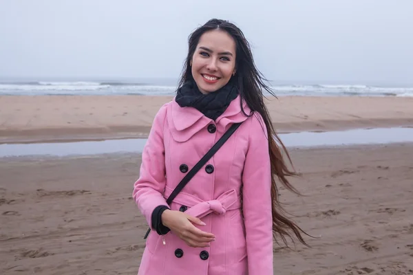 Portrait of attractive young woman with long beautiful hairs dressed pink coat, which posing in autumn North sea beach.