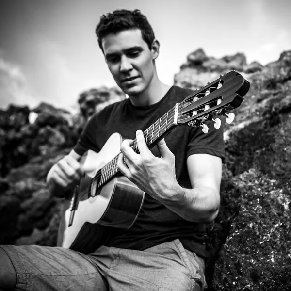 Handsome smiling guitarist play music siting on beach rock. Black-white outdoor photo.