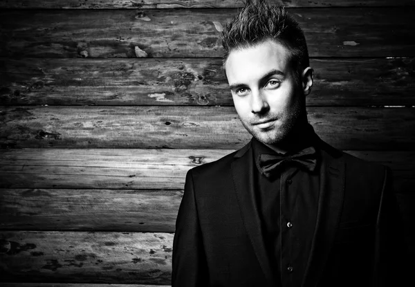 Portrait of young beautiful fashionable man against wooden wall In black suit & bow tie. Black-white fashion photo against wooden background.