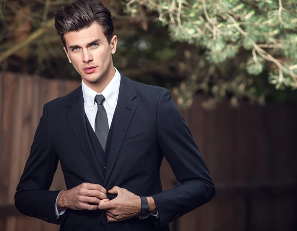 Elegant handsome man in classical suit poses near wooden fence