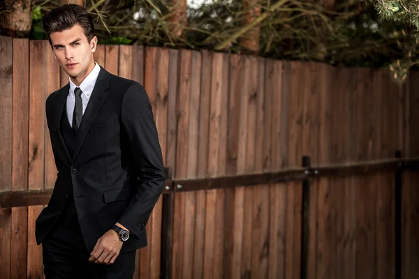 Elegant handsome man in classical suit poses near wooden fence