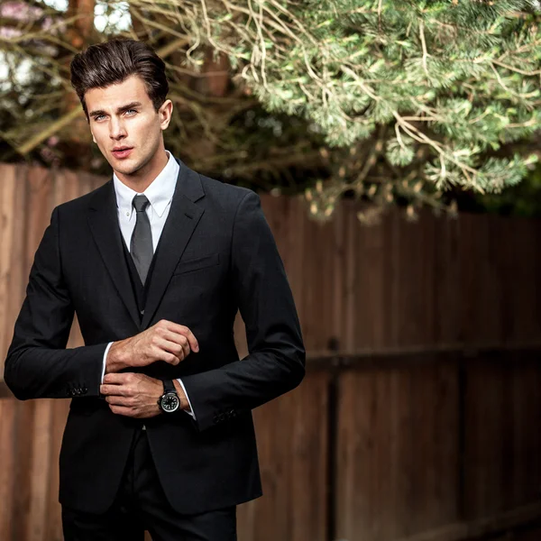 Elegant handsome man in classical suit poses near wooden fence