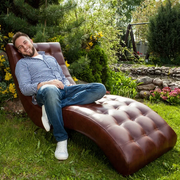 Young handsome man in casual clothes sit in luxury sofa in summer garden.