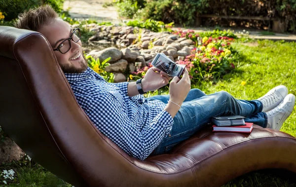 Young handsome man in casual clothes & glasses sit in luxury sofa in summer garden.