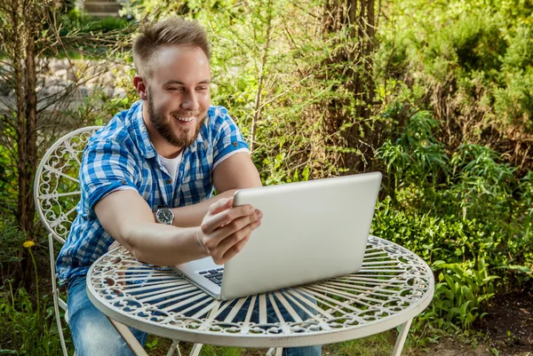 Young smiling handsome man in casual clothes work at an iron table with computer against country garden.