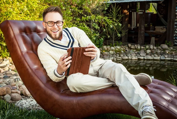 Young handsome man in glasses sit in luxury sofa with iPad in summer garden.