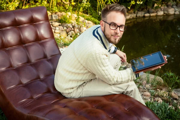 Young handsome man in glasses sit in luxury sofa with iPad in summer garden.