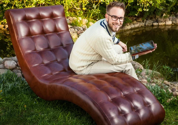 Young handsome man in glasses sit in luxury sofa with iPad in summer garden.