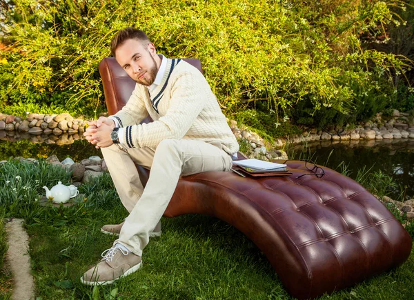 Young handsome man in glasses sit in luxury sofa with iPad in summer garden.