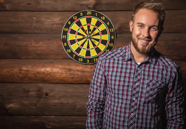 Portrait of young friendly lucky man against old wooden wall with darts game. Concept: Hit in purpose. Photo.