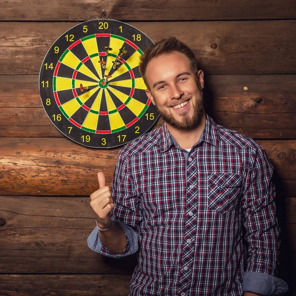 Portrait of young friendly lucky man against old wooden wall with darts game. Concept: Hit in purpose. Photo.