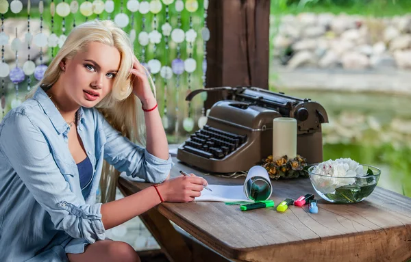 Beautiful young blonde sits in arbor at oak table near vintage typewriter.