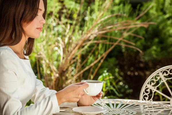 Outdoors portrait of beautiful young girl in luxury white dress posing near garden metal table with cup of coffee.