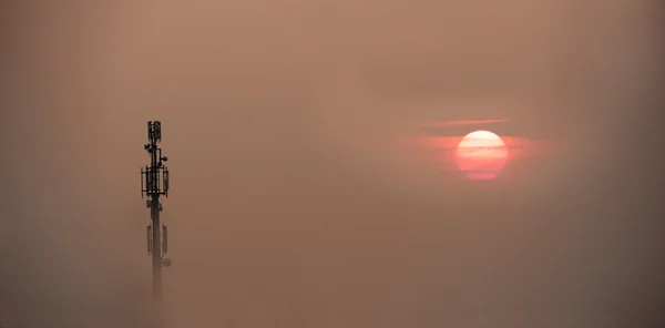 Mobile phone antenna tower emerges from the fog over a sunset sk