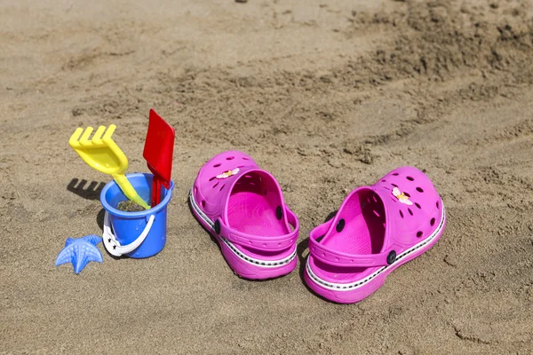 Pink beach crocs and blue sand toys on sandy beach.Beach flip flops in the foreground and blurred sea in the background