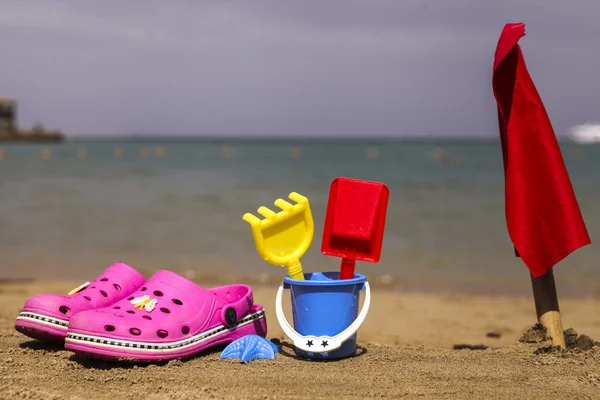 Pink beach crocs and blue sand toys on sandy beach.Beach flip flops in the foreground and blurred sea in the background