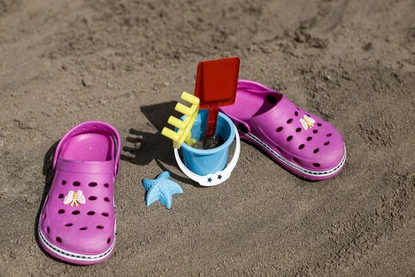 Pink beach crocs and blue sand toys on sandy beach.Beach flip flops in the foreground and blurred sea in the background