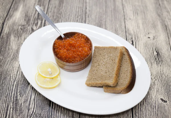Fresh appetizing red salmon caviar in a wooden jar with pieces of bread on a porcelain platter.