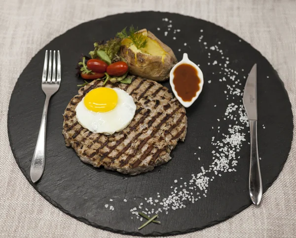 Big juicy grilled steak marbled beef with egg baked potatoes with barbecue sauce. Served on a stone plate with a fork and spoon on a textile background.