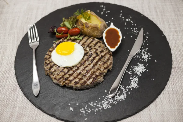 Big juicy grilled steak marbled beef with egg baked potatoes with barbecue sauce. Served on a stone plate with a fork and spoon on a textile background.