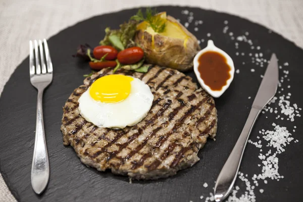 Big juicy grilled steak marbled beef with egg baked potatoes with barbecue sauce. Served on a stone plate with a fork and spoon on a textile background.