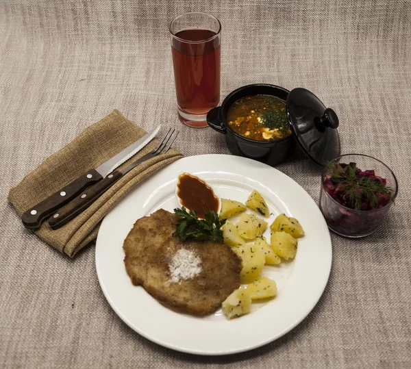 Business lunch : Petite Wiener schnitzel with boiled potatoes and ketchup. Served on a white porcelain plate with fork and knife on a wooden background. Beet  salad and tomato, red pepper soup, sauce