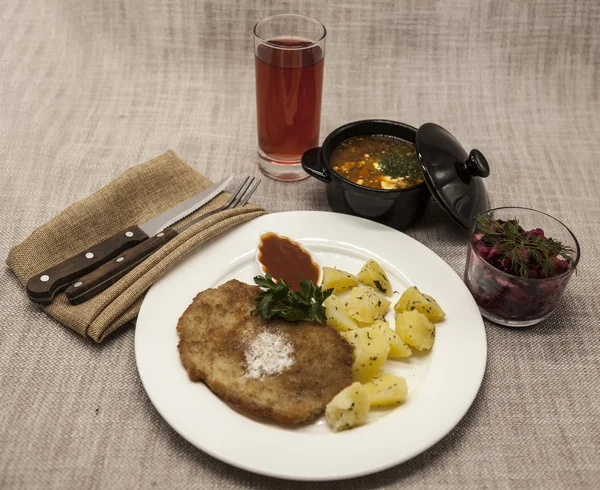 Business lunch : Petite Wiener schnitzel with boiled potatoes and ketchup. Served on a white porcelain plate with fork and knife on a wooden background. Beet  salad and tomato, red pepper soup, sauce