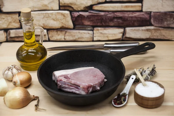 A piece of delicious fresh raw pork close-up on a cast-iron frying pan, onions, garlic, spices, salt, olive oil, fork, knife on a rustic kitchen table