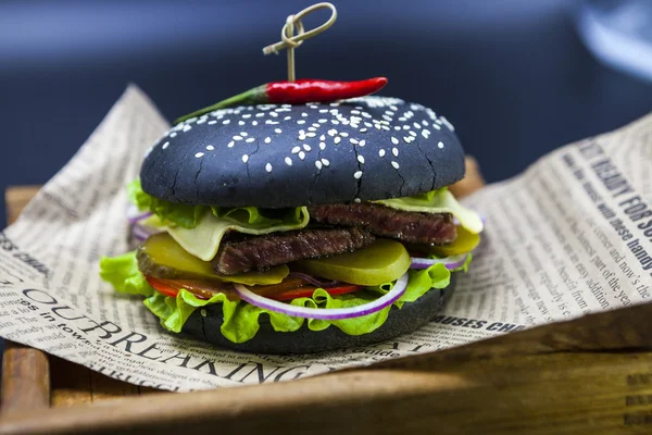 Black burger. A burger with a black roll slices of juicy marble beef, fused cheese, fresh salad and sauce of a barbecue. A burger on the newspaper on a wooden tray  on a dark background.