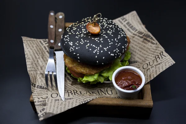 Black burger. A burger with a black roll slices of juicy marble beef, fused cheese, fresh salad and sauce of a barbecue. A burger on the newspaper on a wooden tray with a knife and a fork on a dark ba