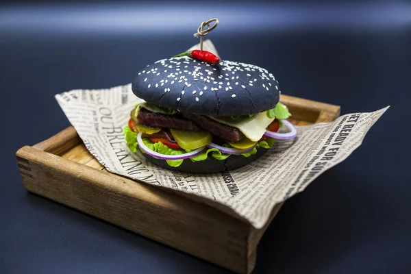 Black burger. A burger with a black roll slices of juicy marble beef, fused cheese, fresh salad and sauce of a barbecue. A burger on the newspaper on a wooden tray  on a dark background.