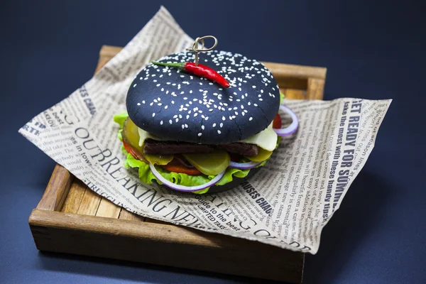 Black burger. A burger with a black roll slices of juicy marble beef, fused cheese, fresh salad and sauce of a barbecue. A burger on the newspaper on a wooden tray  on a dark background.