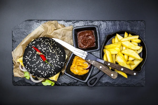 Set of the black burger. Stone plate with: black burger roll slices of juicy marble beef, fused cheese, fresh salad with French fries on a cast-iron griddle, with chili sauce and guakomole.