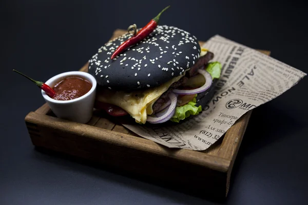 Black burger. A burger with a black roll slices of juicy marble beef, fused cheese, fresh salad and sauce of a barbecue. A burger on the newspaper on a wooden tray with a knife and a fork on a dark ba