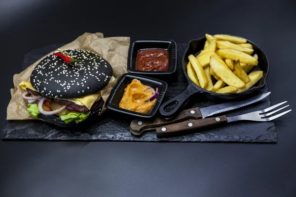 Set of the black burger. Stone plate with: black burger roll slices of juicy marble beef, fused cheese, fresh salad with French fries on a cast-iron griddle, with chili sauce and guakomole.