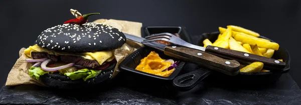 Set of the black burger. Stone plate with: black burger roll slices of juicy marble beef, fused cheese, fresh salad with French fries on a cast-iron griddle, with chili sauce and guakomole.