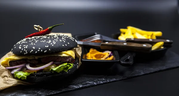 Set of the black burger. Stone plate with: black burger roll slices of juicy marble beef, fused cheese, fresh salad with French fries on a cast-iron griddle, with chili sauce and guakomole.