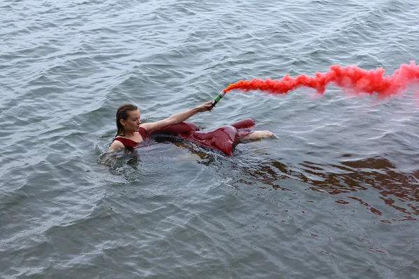 Young beautiful drowned woman with smoke grenade in red dress floating in the water after ship crush.